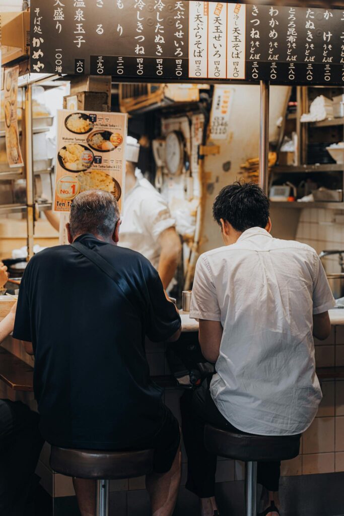 Two men sitting at a restaurant bar enjoying authentic Asian cuisine with a chef working in the background.